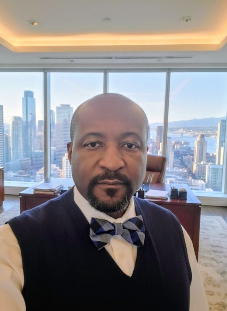 Man in a bow tie posing indoors with a city skyline behind.